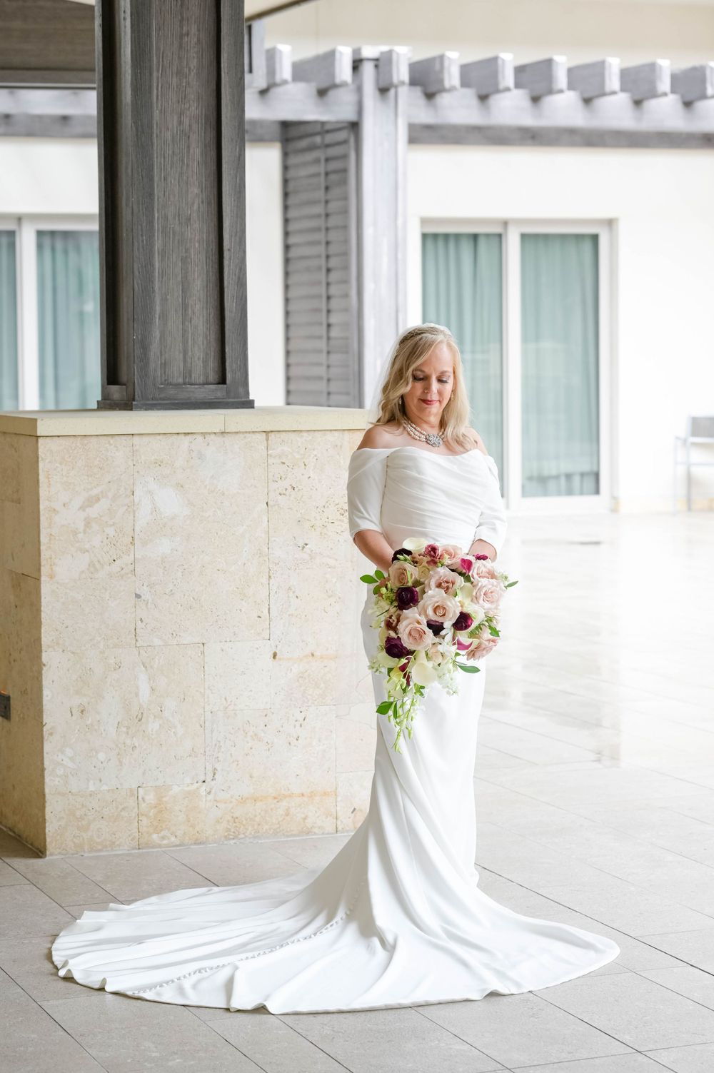 A bride in a white off-shoulder gown with train holds a colorful bouquet on a covered patio.