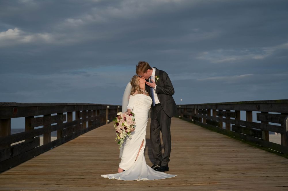 Romantic couple shares an intimate moment on a wooden pier against dramatic cloudy skies.