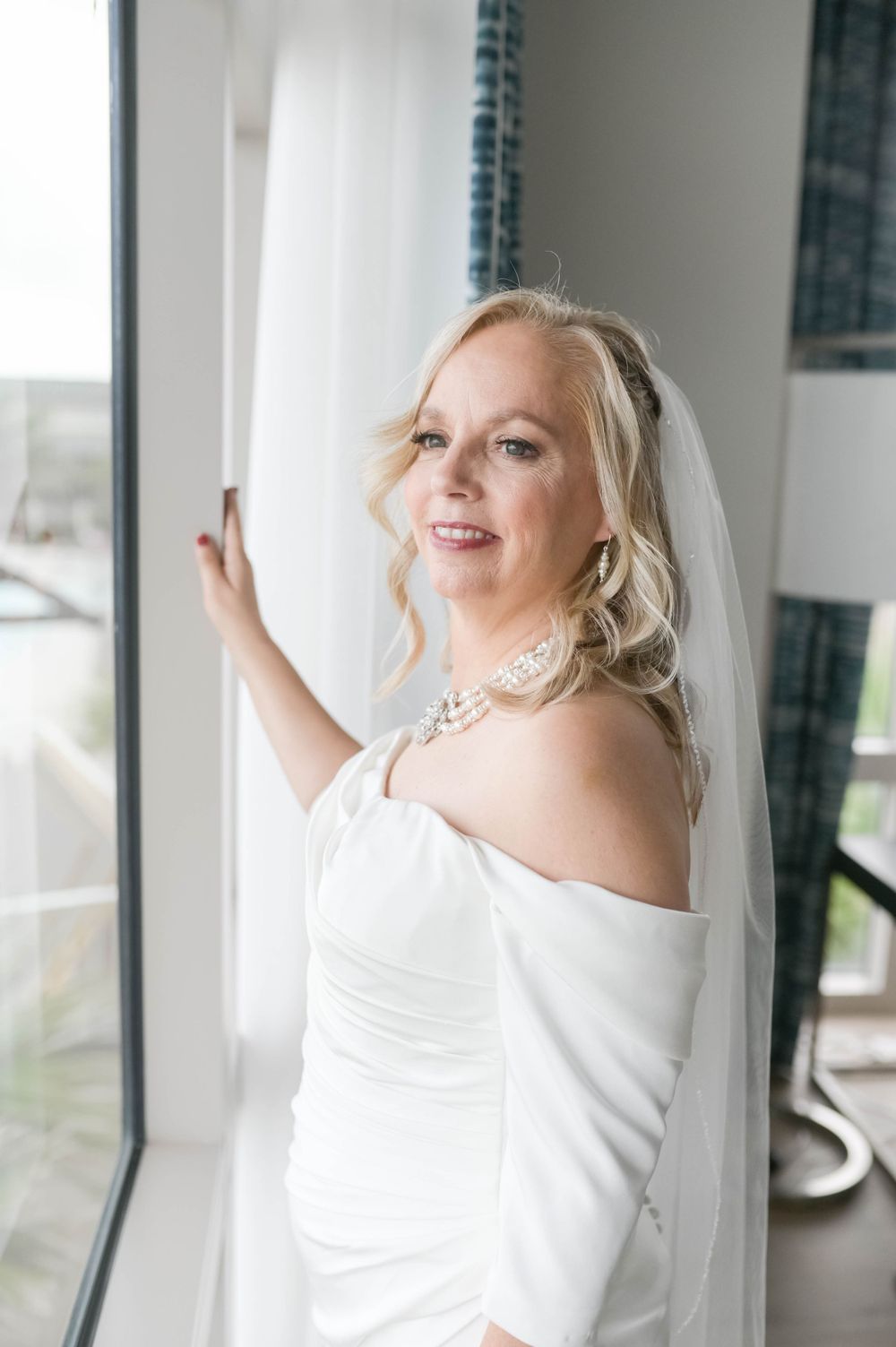 A bride in an off-shoulder white wedding dress stands by a window in natural light.