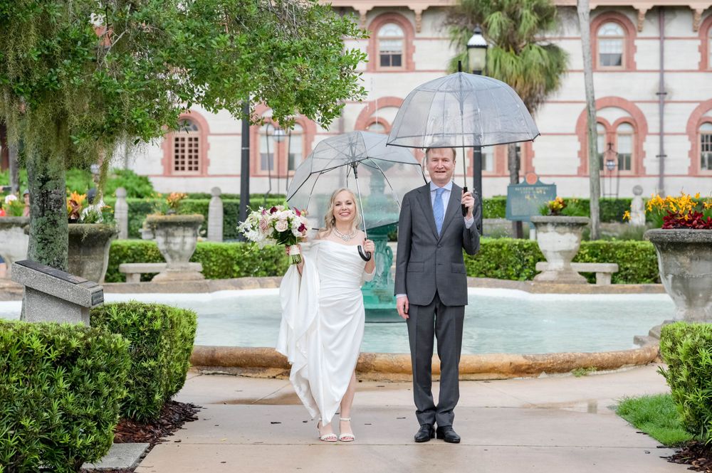 A couple poses with an umbrella in front of a grand historic building on their wedding day.