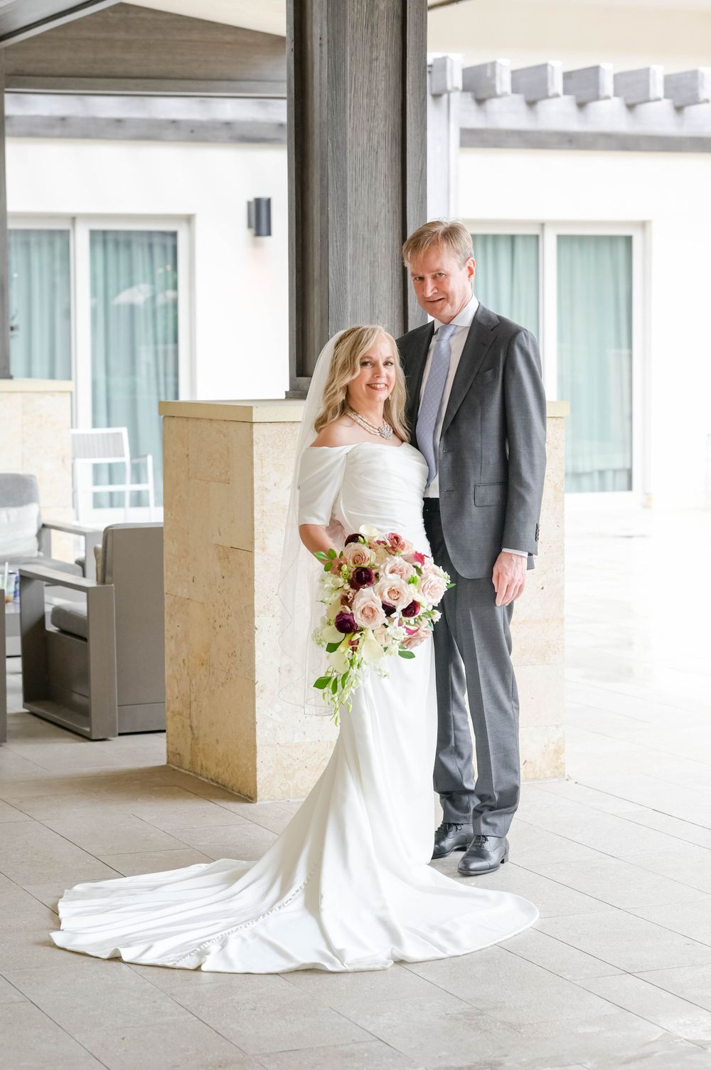 A newlywed couple poses on a covered patio in formal wedding attire.