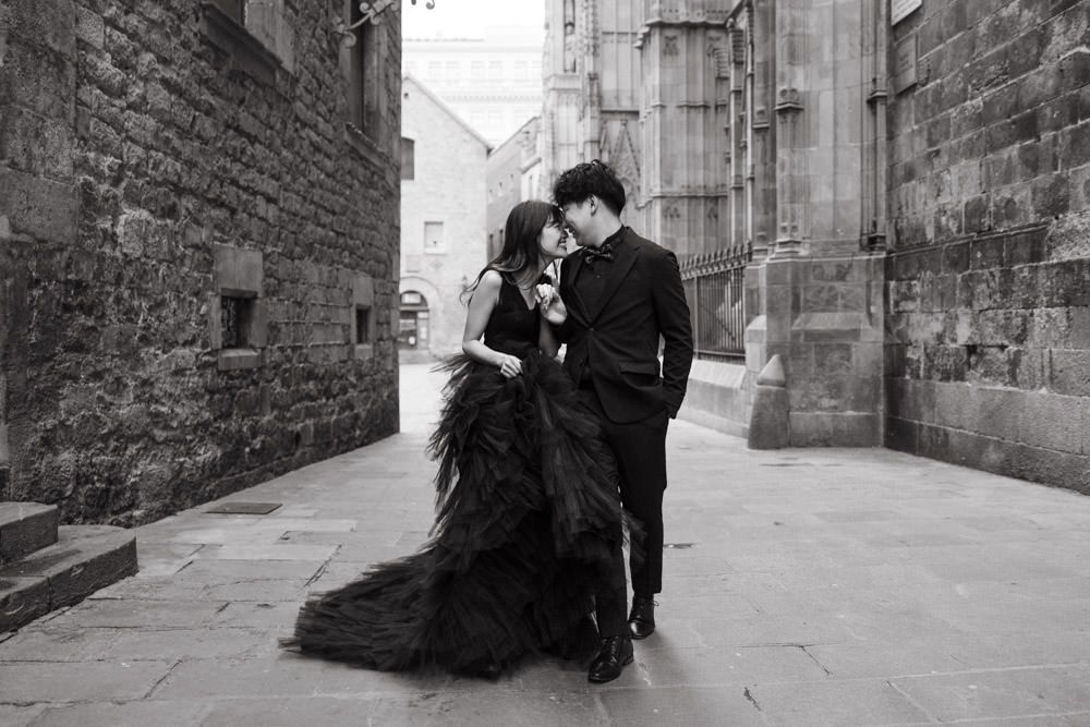A couple in formal black attire poses against historic stone architecture.