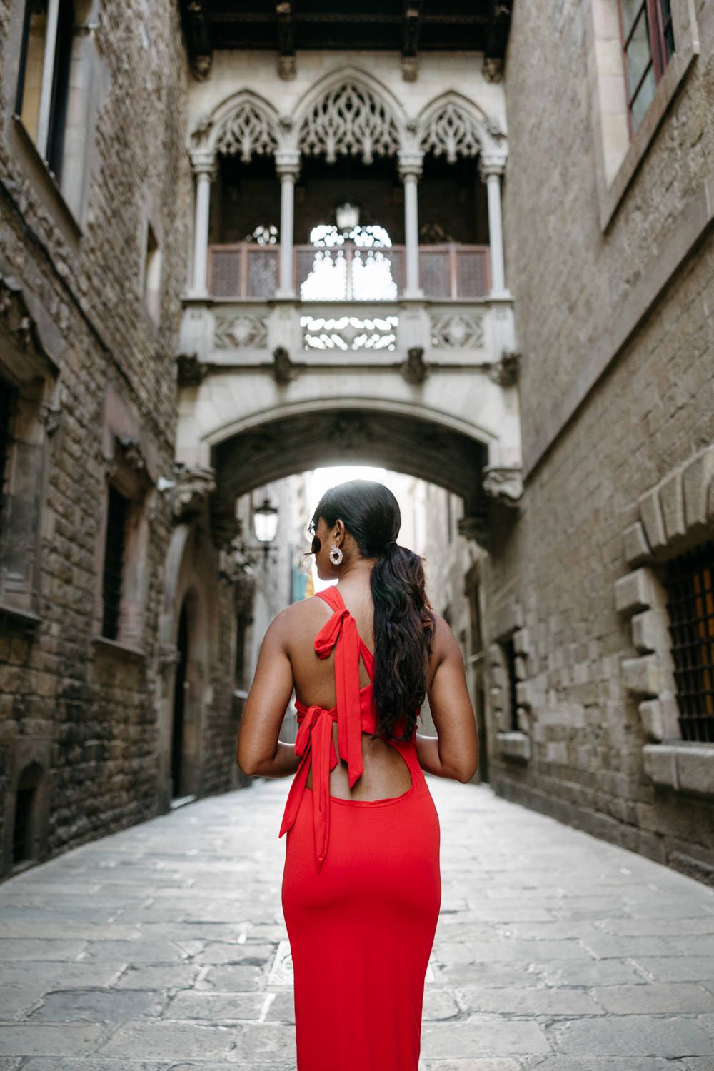 Woman in red dress walking through historic Gothic archways and stone corridors in European city.