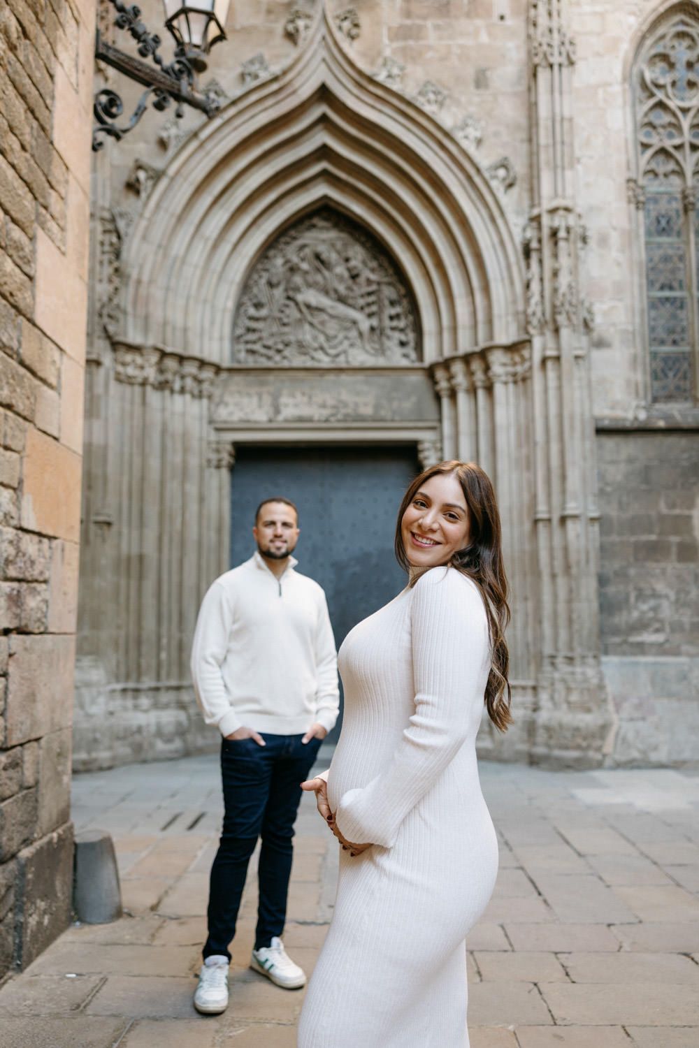 Couple in white and dark outfits posing playfully in front of ornate Gothic cathedral entrance.