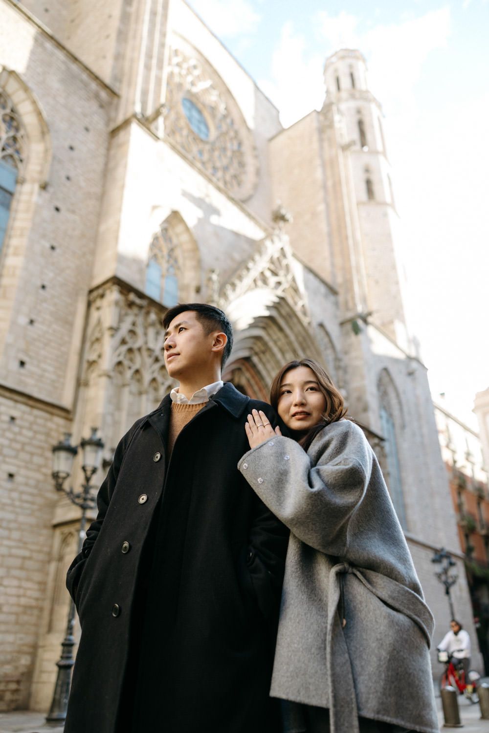 A stylish couple in winter coats poses in front of a Gothic cathedral's ornate facade.