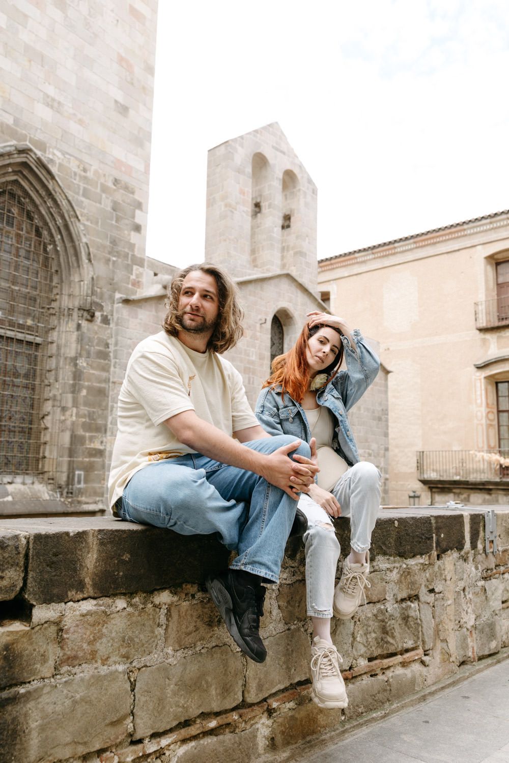 A couple in casual denim outfits sits on a stone wall with a historic church bell tower in the background.