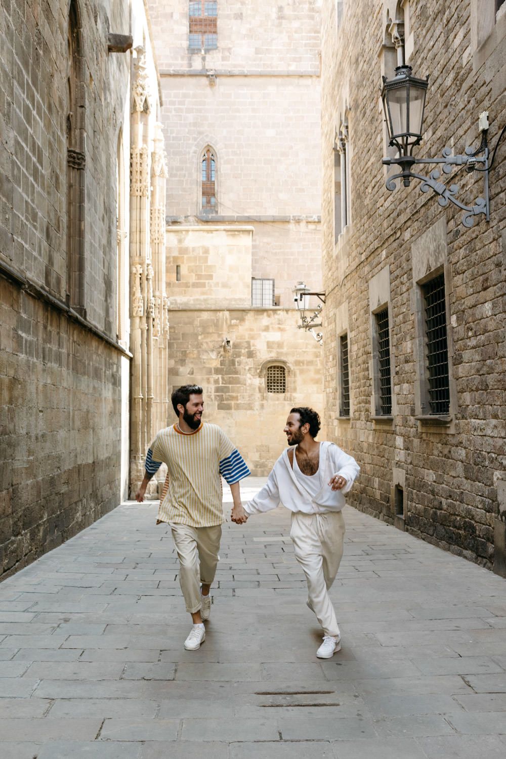 Two friends running and laughing through narrow medieval stone streets in casual summer outfits.
