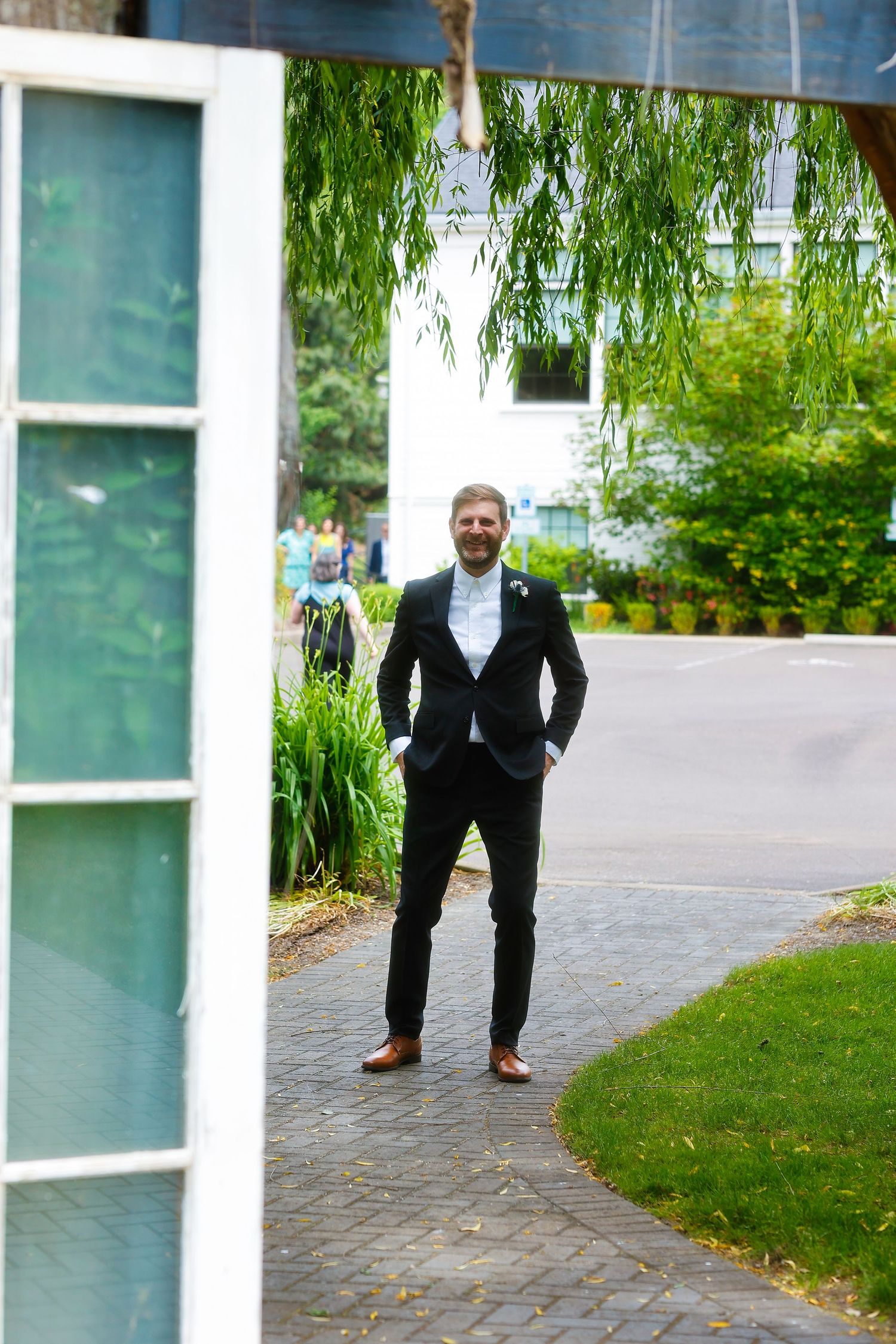 Groom in black suit standing confidently on a stone path with greenery and white building in background.