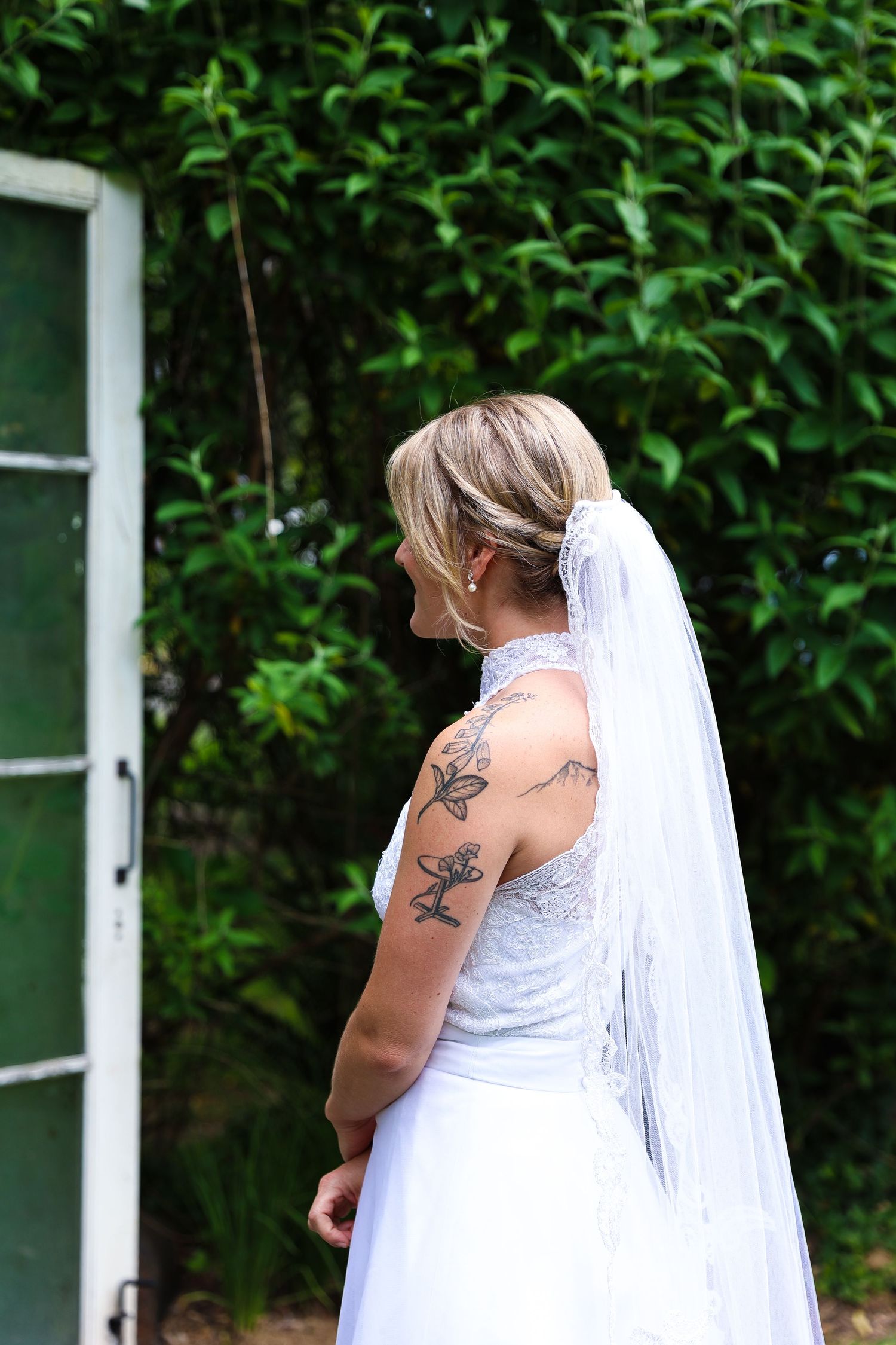 Bride in white dress with tattoos and veil standing near a vintage window frame and greenery.