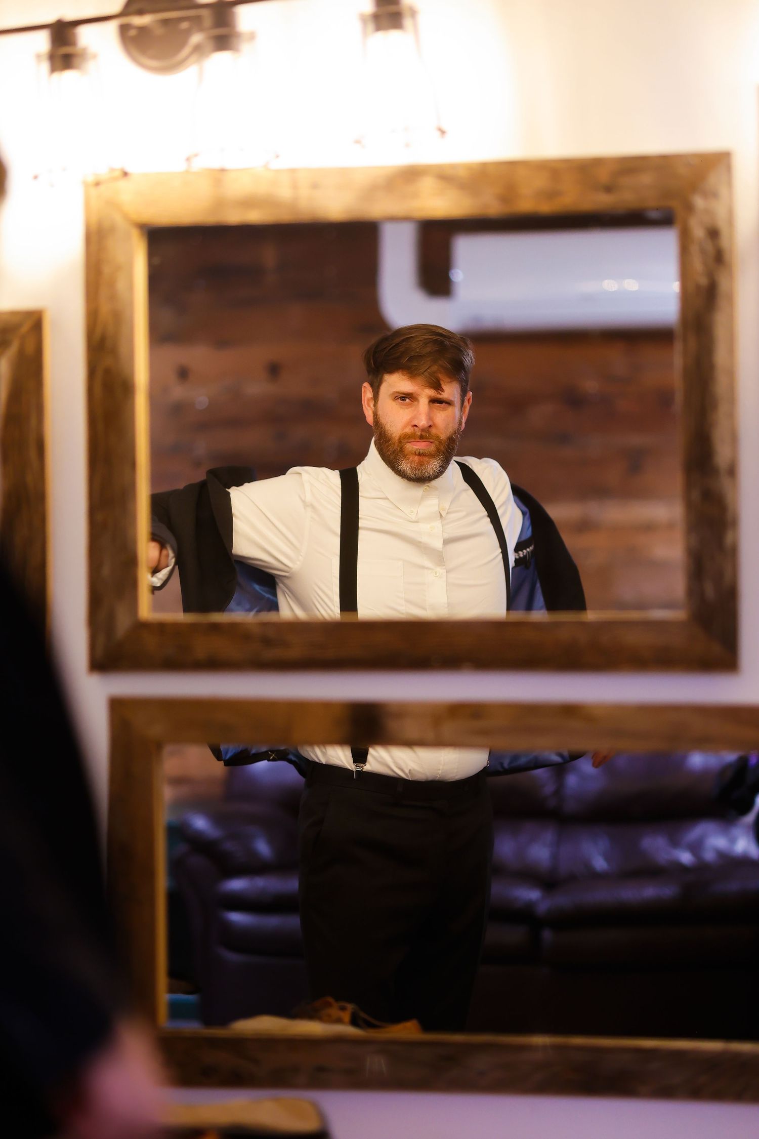 A reflection shows someone adjusting a white dress shirt and suspenders in a wooden framed mirror.