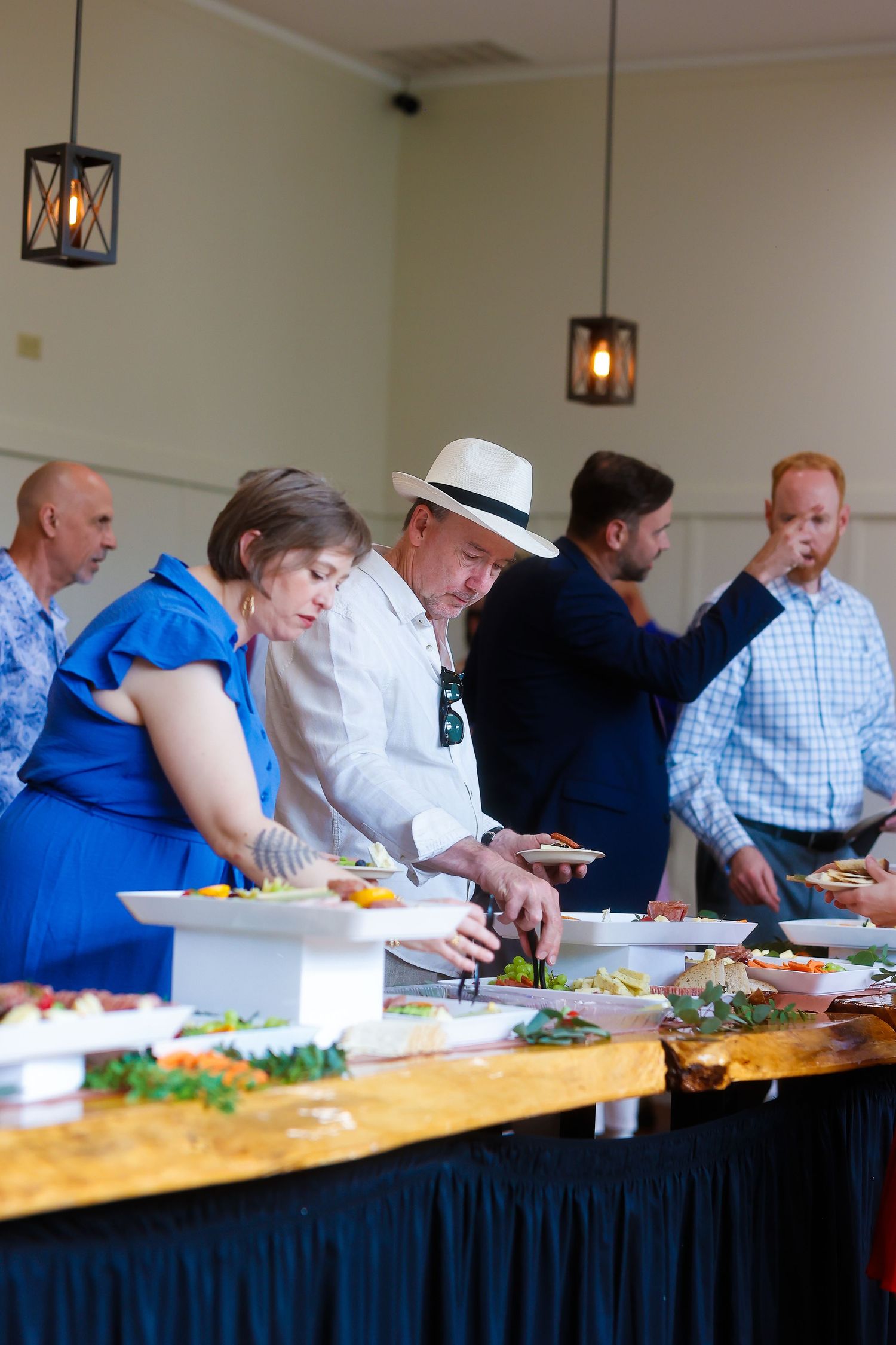 Guests serve themselves from a buffet table decorated with pendant lighting at an indoor event.