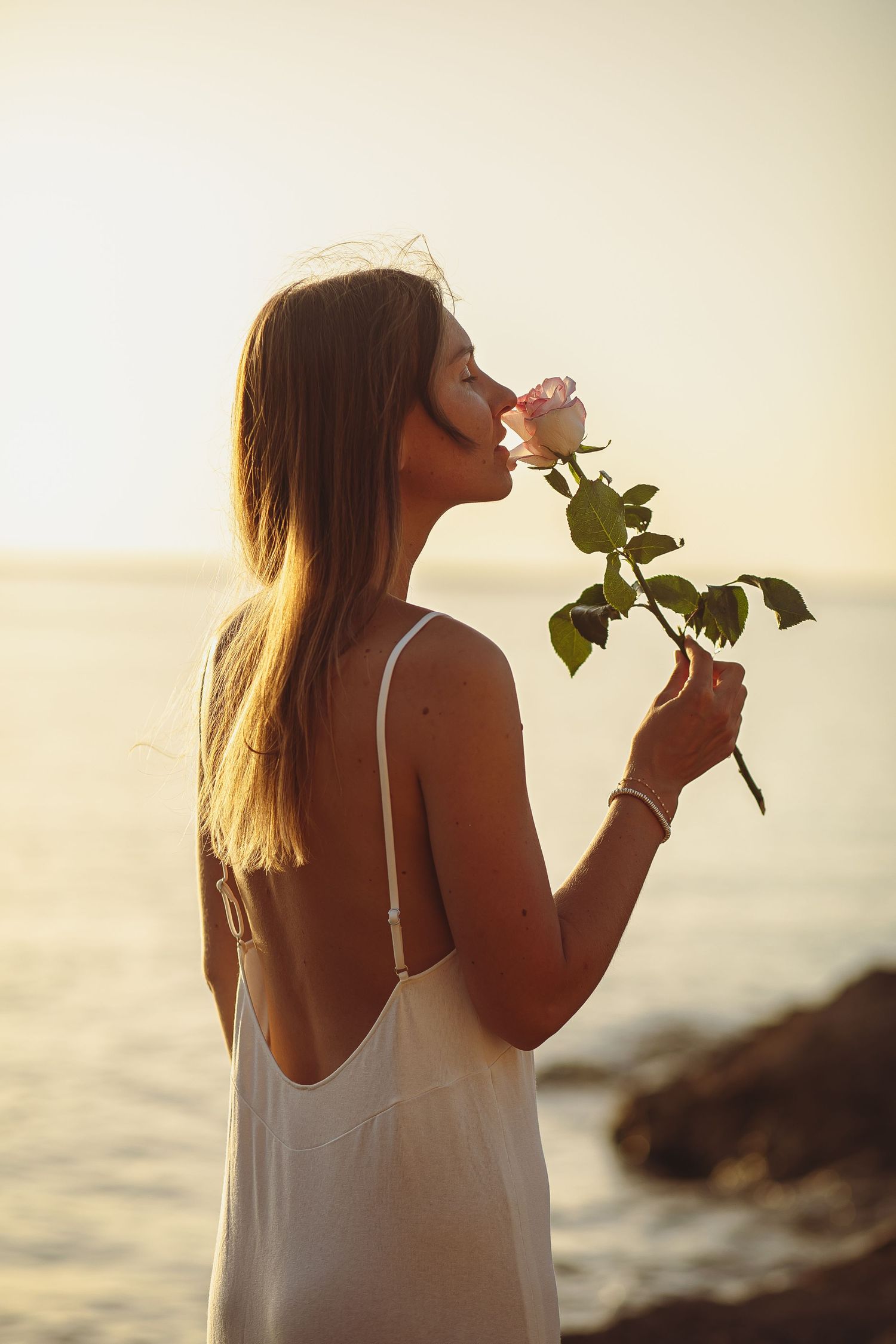 Someone in a white camisole holds a flower against a serene ocean backdrop.