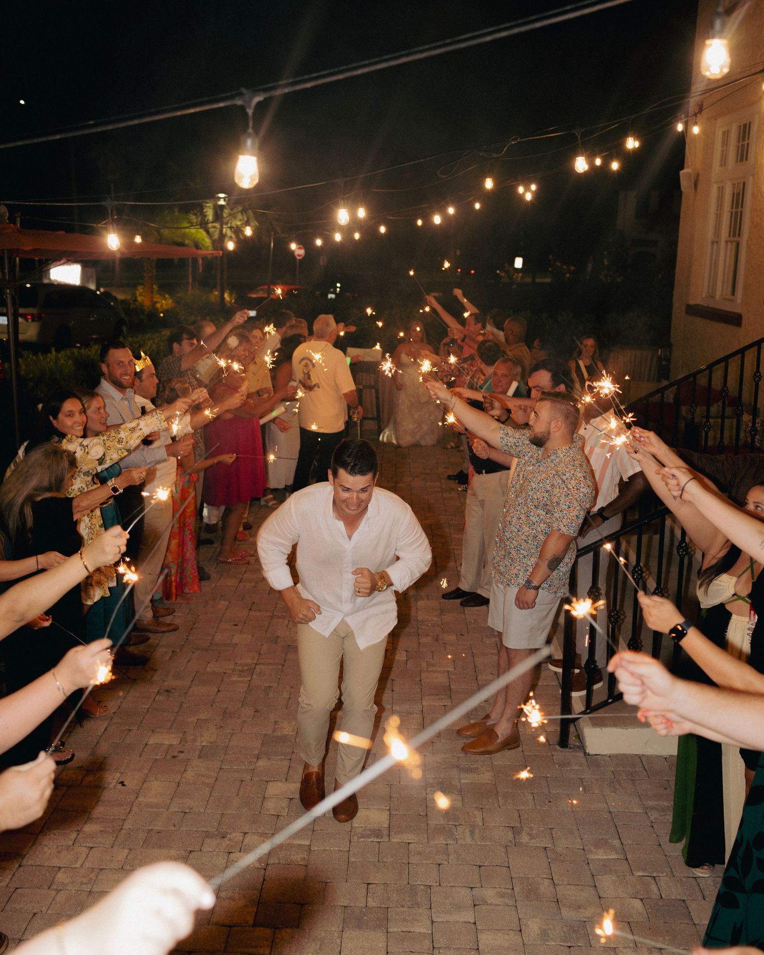 Groom walks through a sparkler tunnel surrounded by wedding guests under string lights at a nighttime reception.