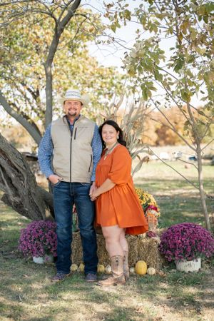 Couple in casual fall attire poses by tree with autumn decorations and purple mums.
