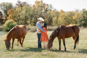 Two brown horses graze in a fall meadow while a couple in country attire stands between them.