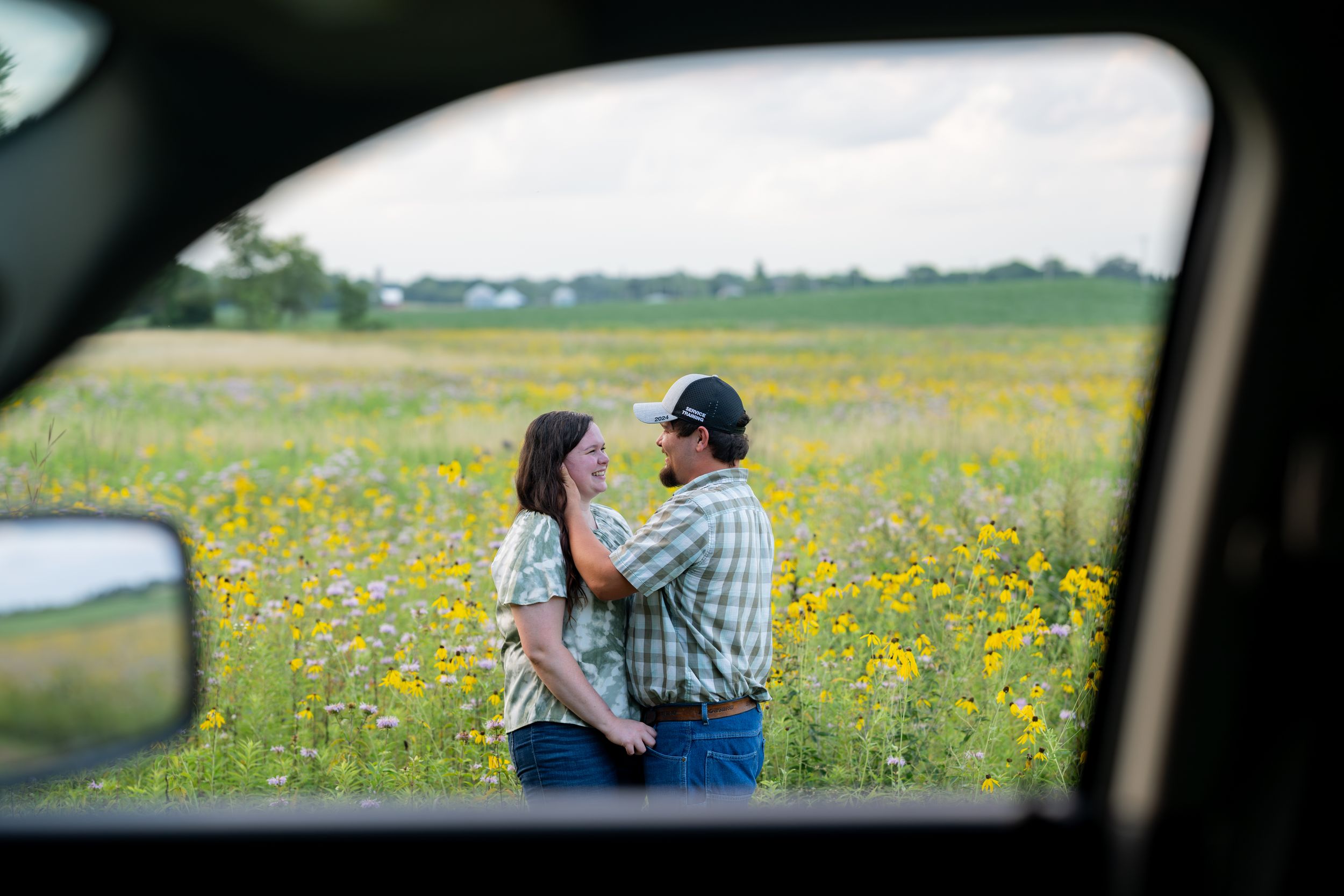Couple embracing in wildflower field viewed through car window frame.