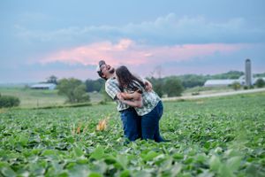 Couple embraces dramatically against sunset sky in rural farm setting.