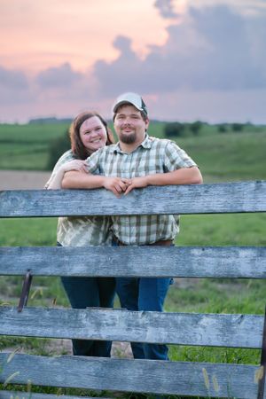A couple enjoys a sunset moment together while leaning on a rustic wooden fence in a pastoral field setting.