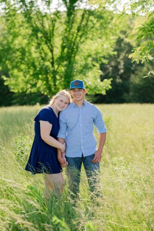 A couple poses together in a sunlit meadow with green trees in the background.