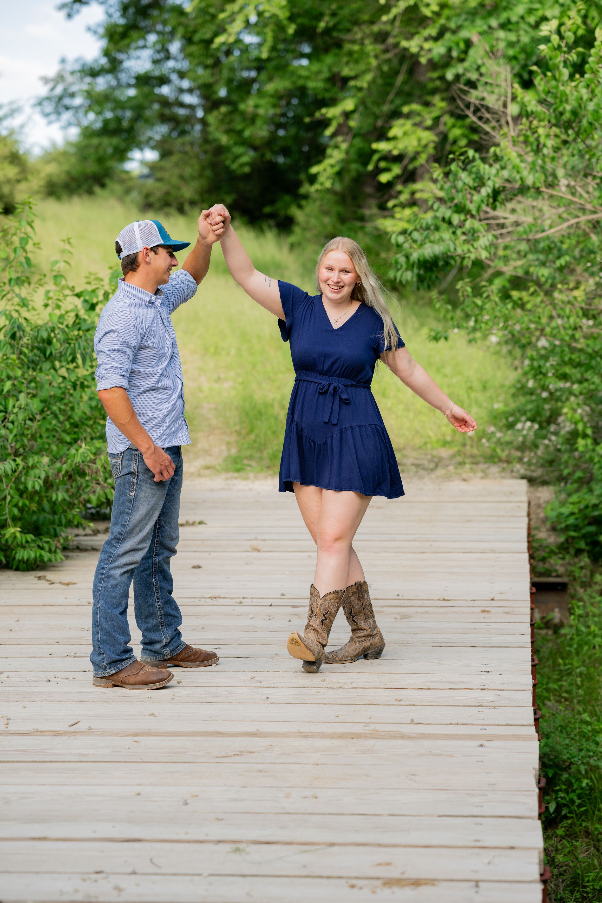 Two people dancing and holding hands on a wooden boardwalk surrounded by lush greenery.