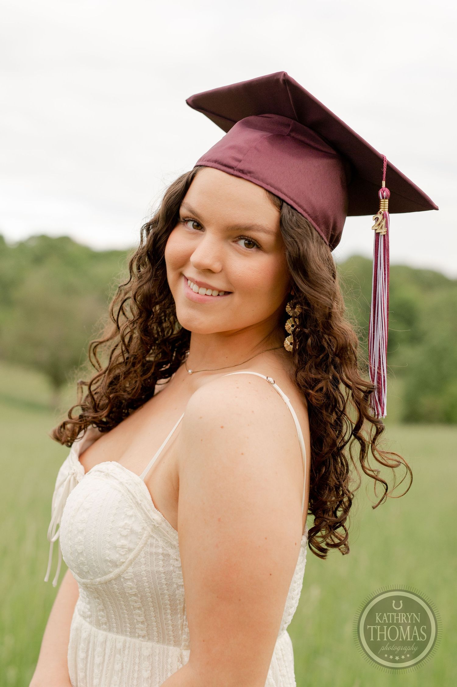 Graduate with curly hair wears maroon cap with tassel and white dress against natural backdrop.
