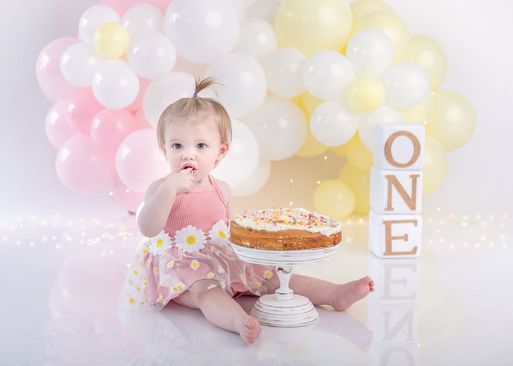 Baby in pink dress reaching for cake with balloon backdrop and ONE letters.