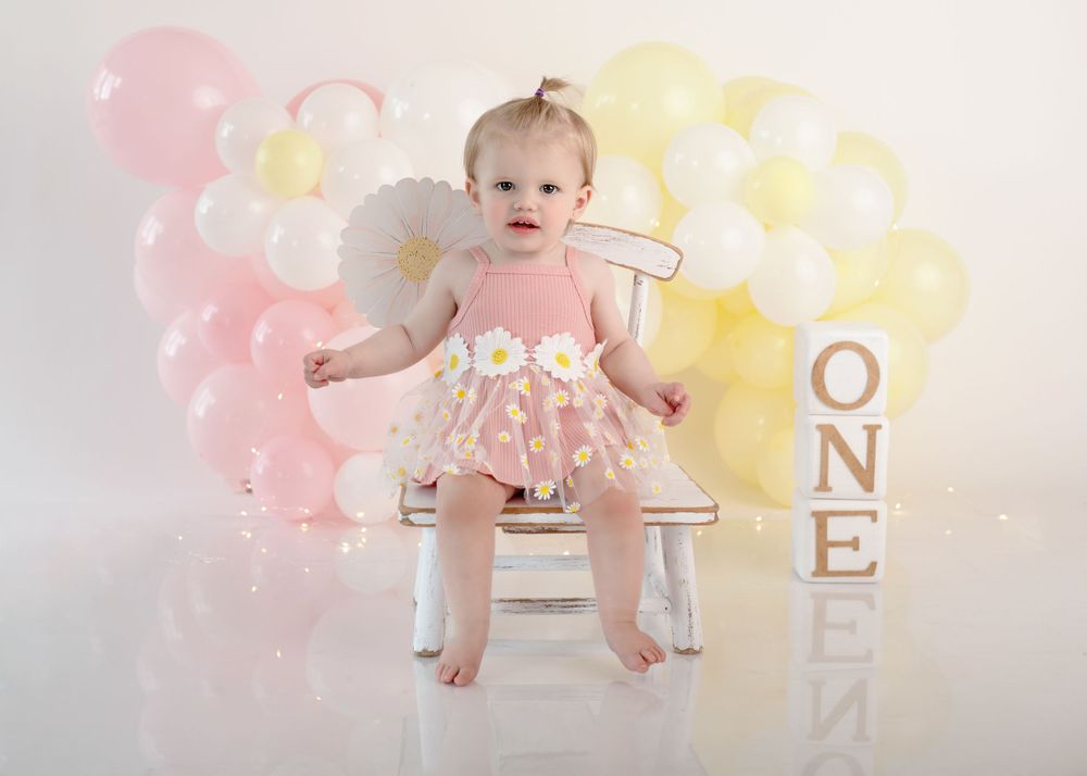 Baby in pink dress seated on white chair with festive balloon background.