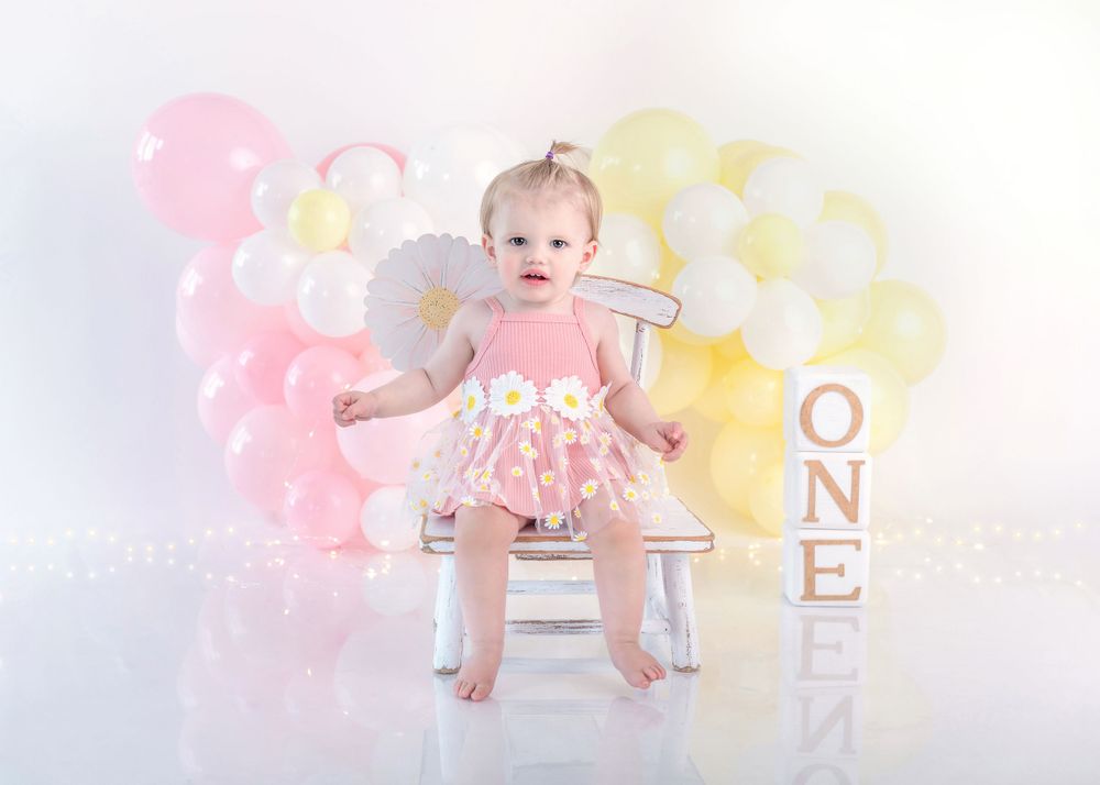 Baby in pink dress sits on chair with pink and yellow balloon backdrop and ONE letters.