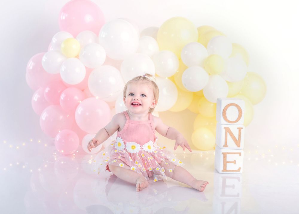 Baby in tulle dress smiles with pink and yellow balloon display and ONE letters.