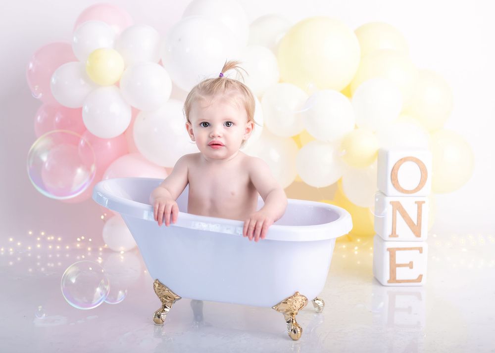 Classic first birthday photo with white miniature bathtub and soft balloon background.