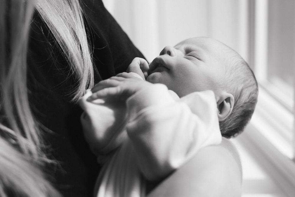 A tender black and white portrait of a newborn baby resting against someone's shoulder in natural window light.