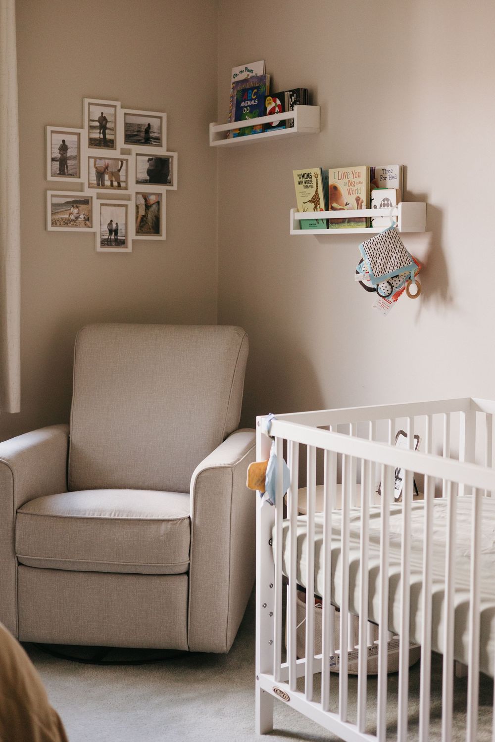 A cozy nursery corner with a grey armchair, white crib, floating shelves and framed photos on the neutral walls.