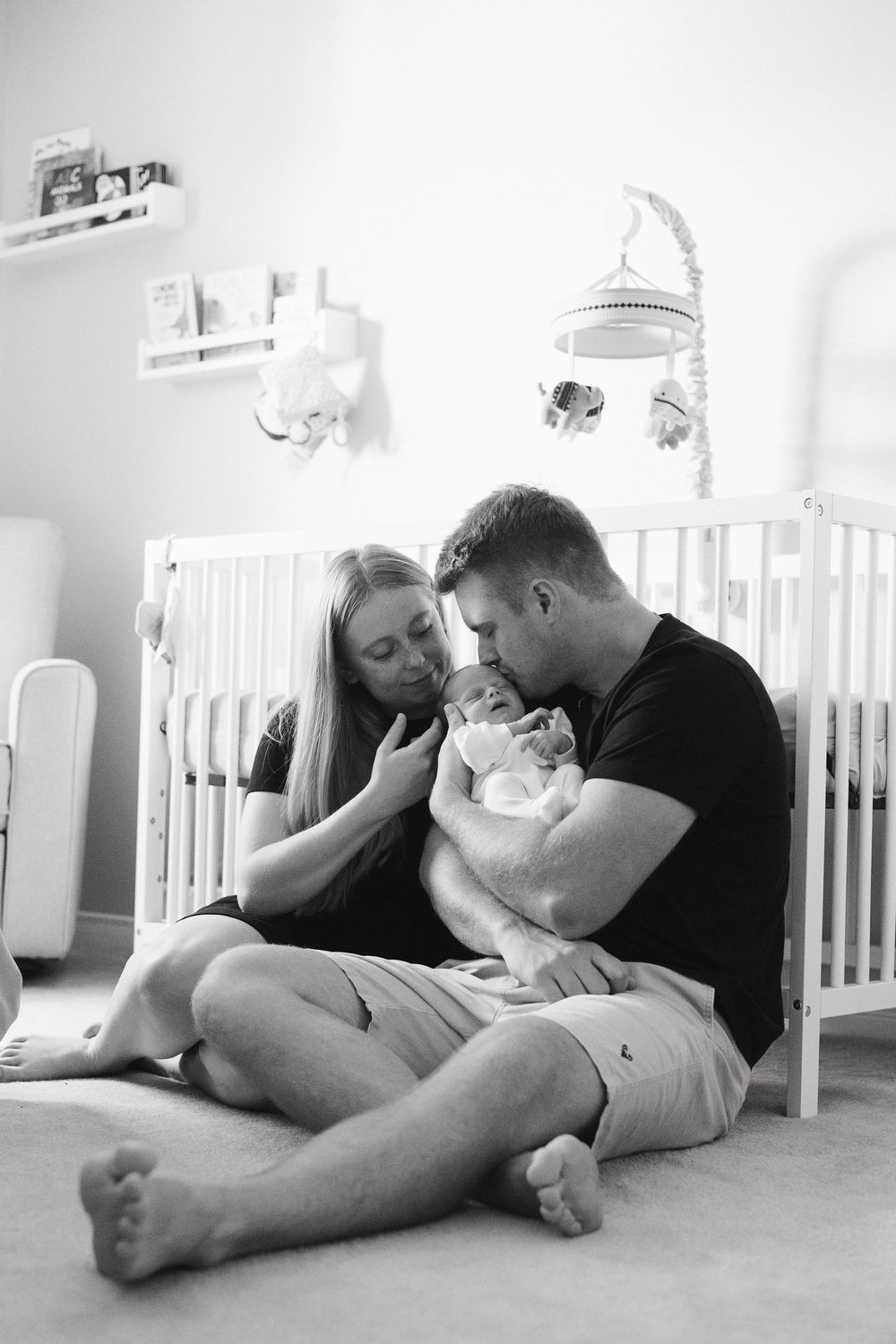 A tender black and white nursery scene with a couple sitting on the floor beside a white crib, cradling their newborn.