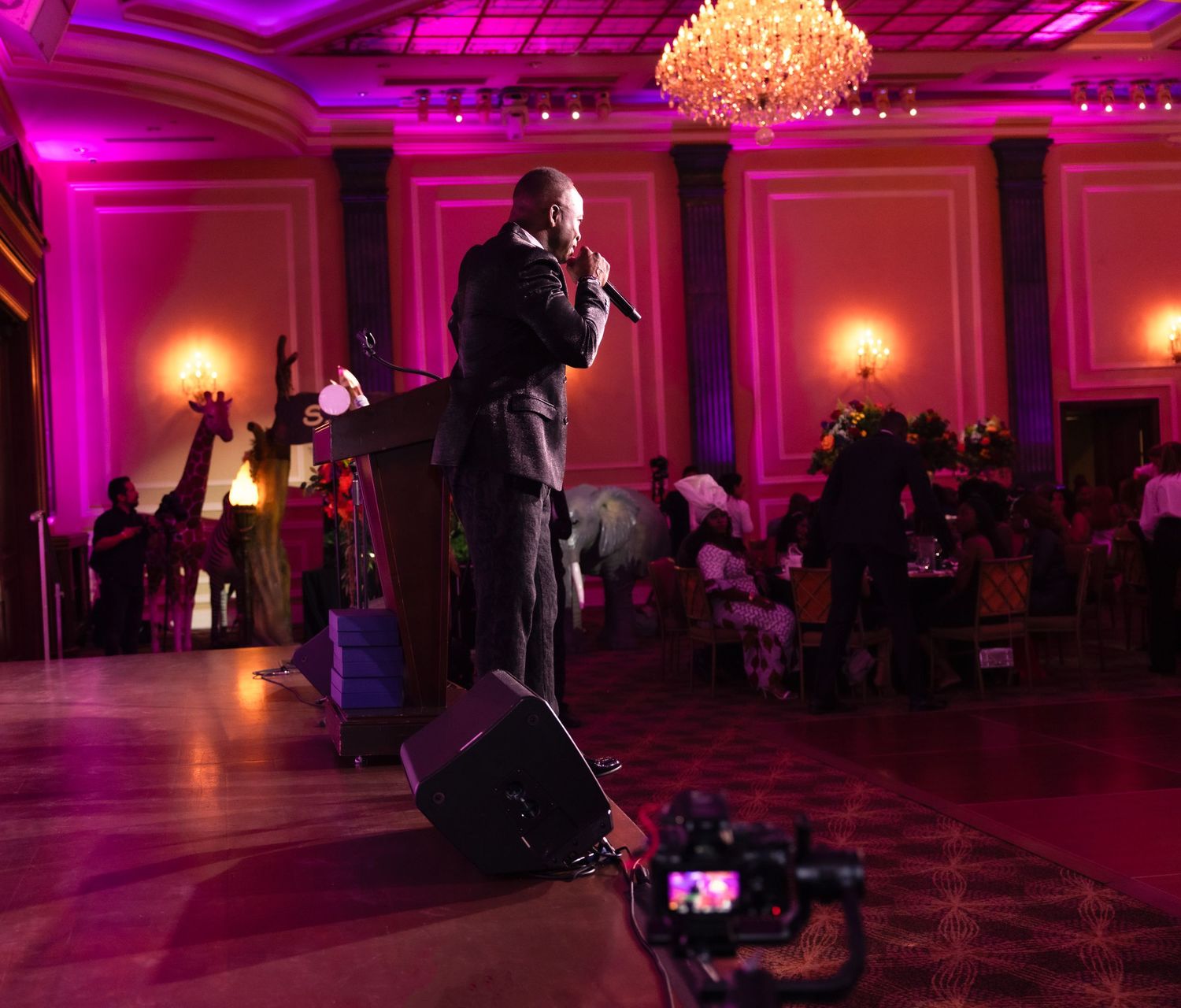A performer on stage with microphone in a ballroom with pink lighting and chandelier overhead.