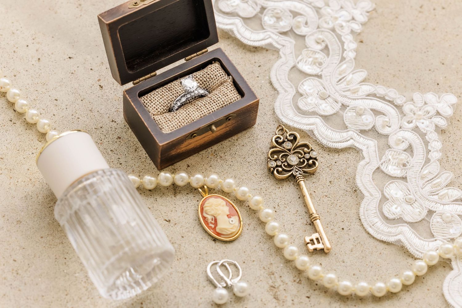 Vintage jewelry and trinkets including a cameo, pearls, lace, perfume bottle and ornate key arranged on a surface.