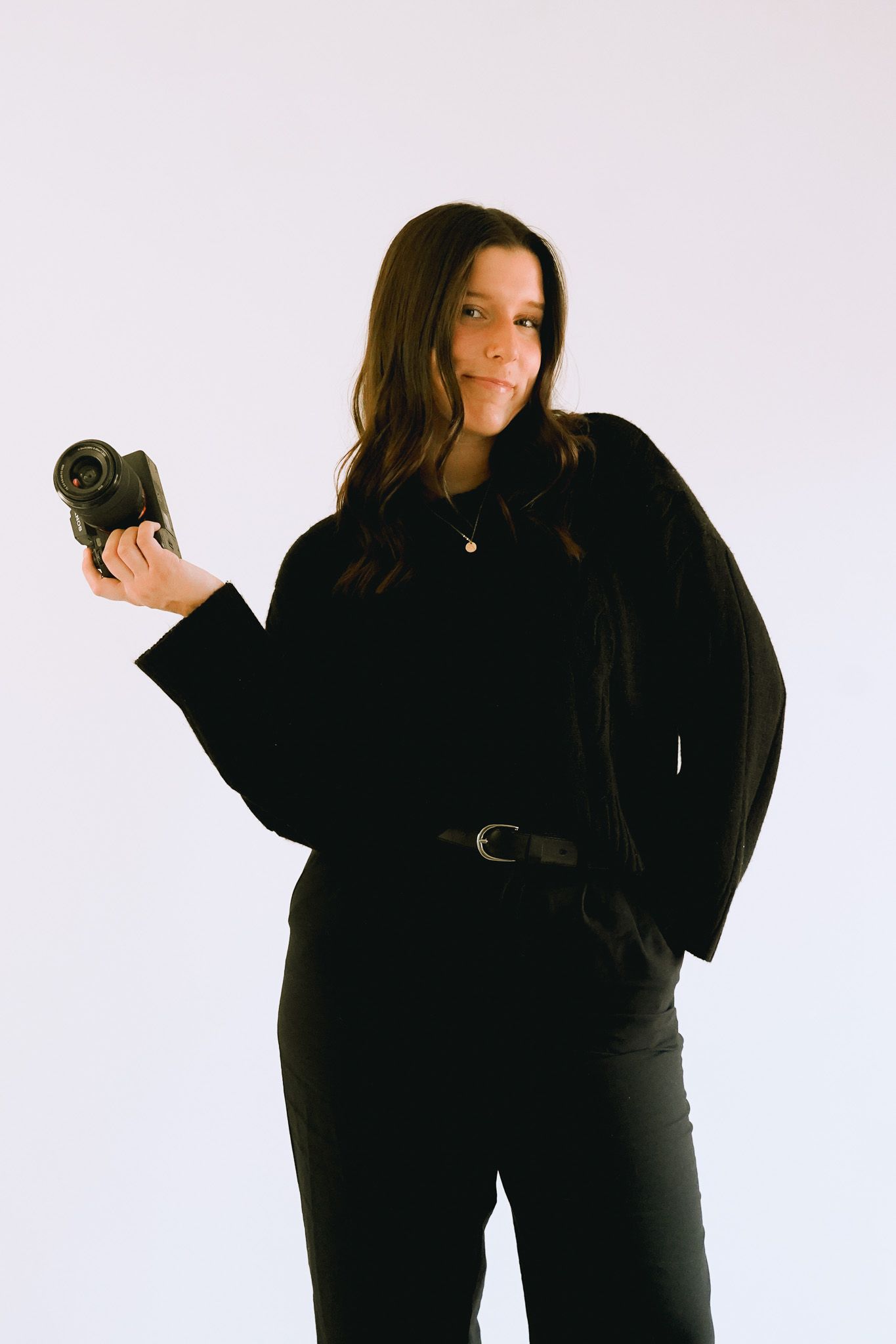 A photographer in all black holds a camera against a light purple studio background.