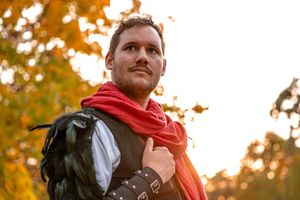 Person wearing a red scarf and medieval costume stands in autumn sunlight with orange foliage in background.
