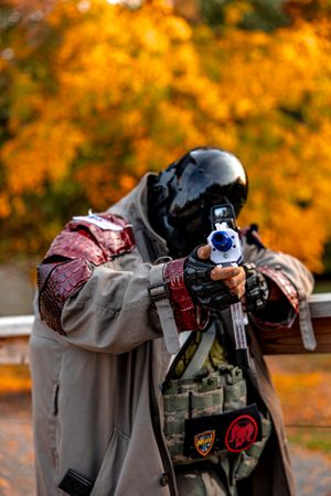 Airsoft player in tactical gear aims rifle against vibrant orange autumn foliage background.