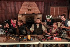 A group of men in vintage attire gathered around a table with antique firearms and bottles in a rustic wooden cabin setting.