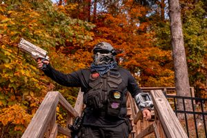 An airsoft player in tactical gear poses with a pistol on wooden stairs against vibrant autumn foliage.