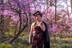 A person in a red dress and black cape stands beneath blooming purple cherry blossom trees in a spring garden.