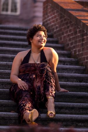 Person in a brown halter dress sits casually on stone steps beside a brick building at sunset.