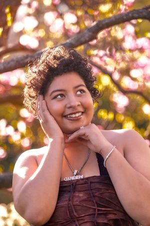 Person in brown dress poses joyfully against blurred pink cherry blossoms in golden sunlight.