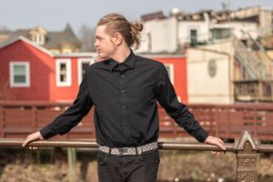 A person in a black button-down shirt leans against a railing with red buildings visible in the background.