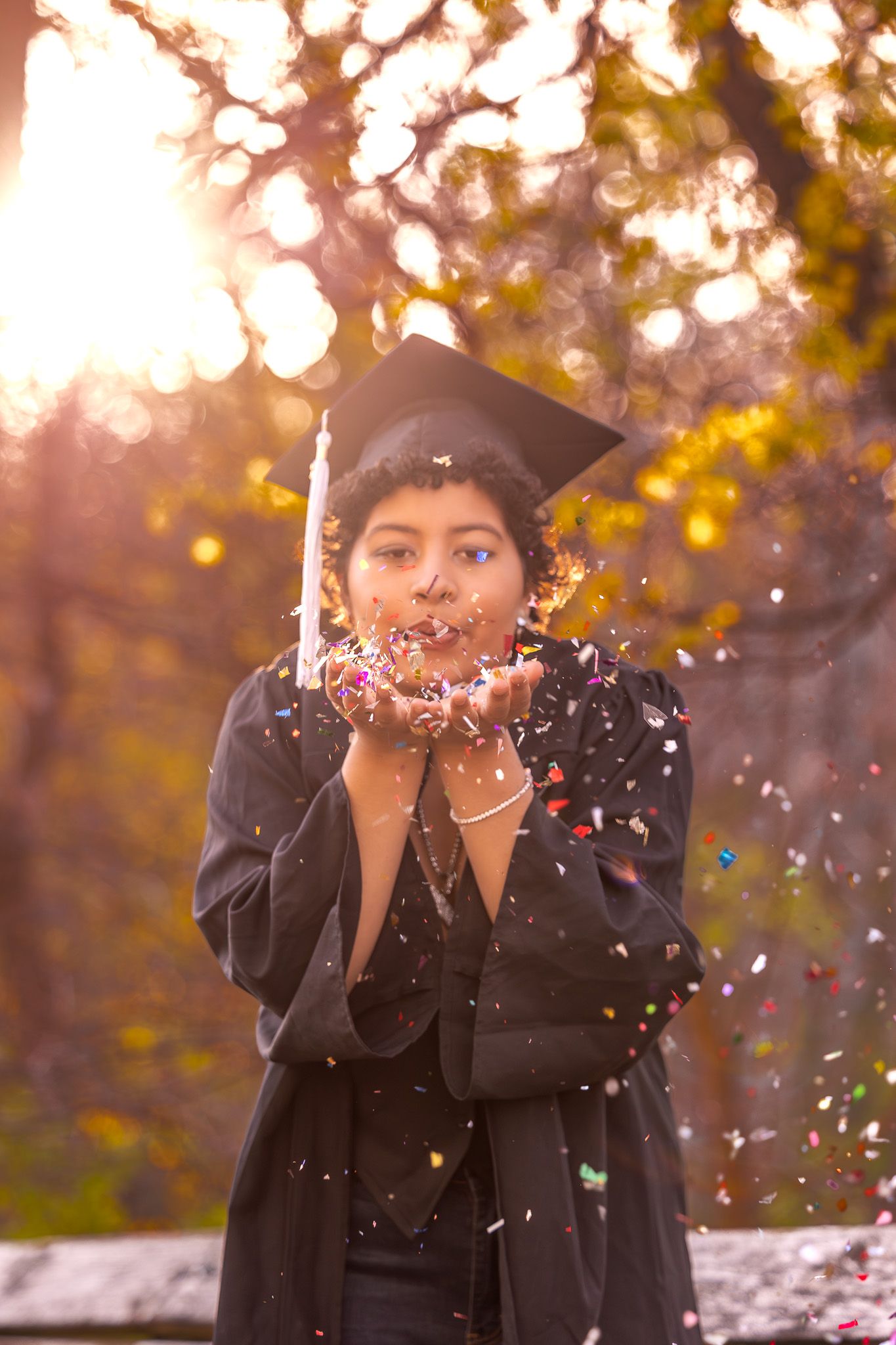 A graduate blows glittery confetti while wearing a cap and gown in warm sunlight.