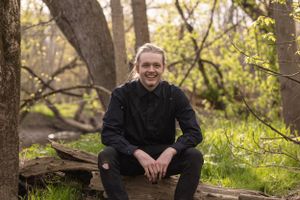 Person in black clothing sits on rustic wooden bench in sunlit wooded setting during autumn season.