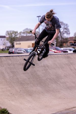 BMX rider performs aerial trick on concrete ramp at skate park with parking lot visible in background.