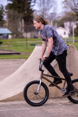 BMX rider in grey shirt and black pants positions their bike on concrete ramp at skate park on cloudy day.