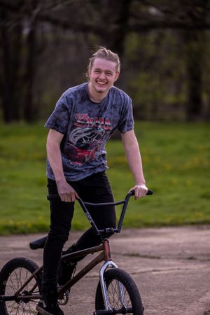 A BMX rider wearing a graphic t-shirt and dark pants smiles while standing with their bike on a dirt path outdoors.