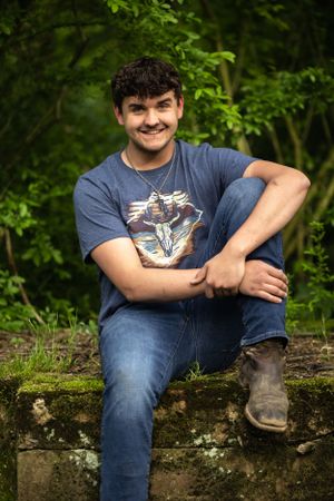 A person sits casually on a stone wall wearing jeans, boots and a t-shirt with a cartoon design in a lush green setting.